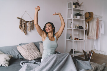 Beautiful young woman smiling while raising hands joyfully in bed, symbolizing gratitude, feminine power and new beginnings in serene, natural bedroom environment.