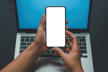 A close-up view of a person holding a smartphone with a blank screen in front of a laptop. Showing concepts of technology and communication