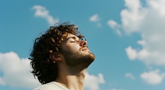 Serene Young Man with Curly Hair Basking in the Sun, Eyes Closed Against a Blue Sky. Adult guy tilts head up and closes eyes in forest and blue sky. He enjoys vacation, meditates, feels calm
