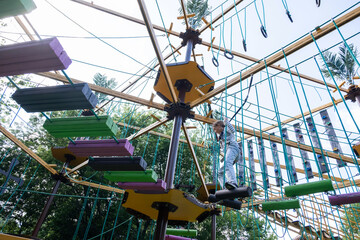 A teenage girl confidently makes her way across a colorful obstacle course in an adventure park, surrounded by trees and enjoying a sunny day. She exhibits balance and focus as she climbs.