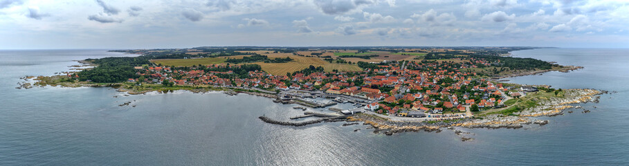 Wide aerial panorama of Svaneke on Bornholm Island Denmark showing red-roofed houses fishing harbor rocky coast and green countryside surrounded by the Baltic Sea under cloudy skies