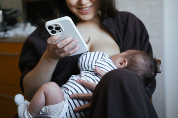 Beautiful young woman is breastfeeding her baby boy at home, close-up view. She is working online at the same time, remote work from home. Chatting with friend, having online shopping. 
