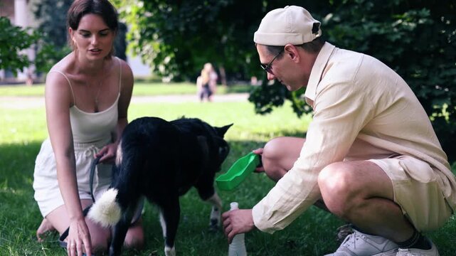 Couple giving water to dog during walking together at pets friendly dog park. Domestic dog with owner enjoy urban outdoor lifestyle on summer vacation. Pet Humanization concept.