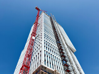 modern high-rise building under construction, viewed from a low angle against a clear blue sky. A large red tower crane stands alongside the building