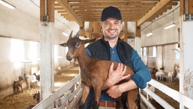 Portrait of handsome Caucasian middle-aged male farmer in cap looking at camera and smiling holding goat in hands in barn. Happy man shepherd caressing cute animal goat at domestic farm