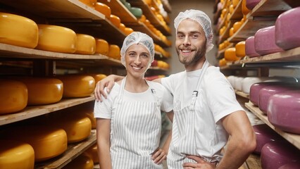 Caucasian young joyful family couple woman and man smiling to camera standing indoor in cheese storage. Dairy products concept. Cheese makers producing cheese at farm house. Portrait shot