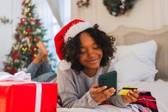 African American woman shopping online holding smartphone paying with gold credit card for Christmas gifts. Girl buying on Internet enter credit card details enjoying Christmas winter holidays at home