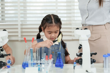 Asian girl holding magnifying glass and observing blue liquid in beaker during elementary science class while standing beside caucasian female teacher at table with colorful test tubes and tools