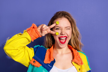 Vibrant young woman showing a playful gesture with smiley stickers, featuring casual and stylish fashion against a vivid purple background.
