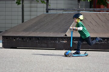 Young boy wearing a black helmet rides a blue three-wheeled scooter at a skate park, concentrating as he moves along the smooth concrete surface. © Karolina