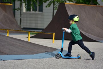 Obraz premium Young boy wearing a black helmet rides a blue three-wheeled scooter at a skate park, concentrating as he moves along the smooth concrete surface.