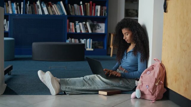 Full shot of biracial female student with pink backpack sitting on floor and studying in university library using laptop and books, writing essay or working on thesis