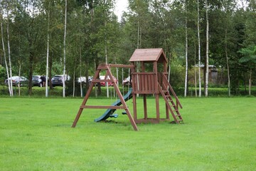 Small wooden playground set with slide and swings on a grassy field near a birch tree grove