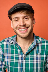 Close-up of cheerful young Caucasian man in flat cap and green plaid shirt, orange background