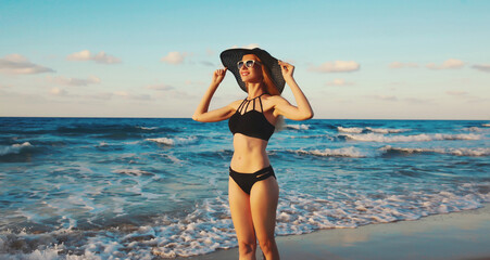 Summer vacation, beautiful happy slim woman in black swimsuit, tourist hat on the beach on sea