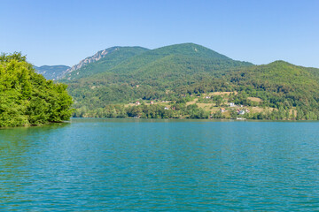 Turquoise water of Perucac Lake with green hills and rural houses on the opposite shore in Bosnia and Herzegovina. Sunny day with blue sky. Tara National Park, Serbia.

