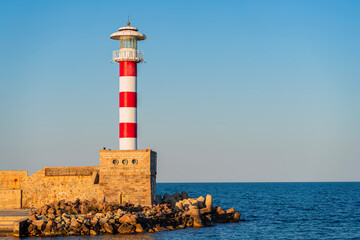 Red and white striped lighthouse on stone pier in Bulgaria Black Sea harbor during golden hour. Concept of coastal tourism, nautical navigation, iconic travel landmark