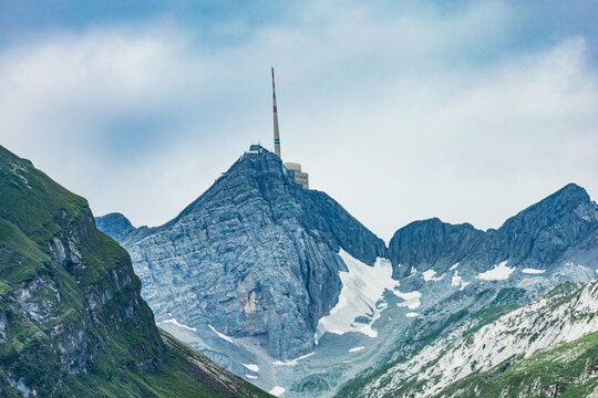 Majestic view of Santis mountain peak in the Swiss Alps, featuring the iconic transmission tower, surrounded by rocky cliffs and patches of snow under a partly cloudy sky.
