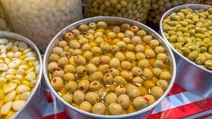 A metal bowl with green olives, some with pits and others stuffed with a red piece of pepper, immersed in a clear liquid. The lighting highlights the color and filling of the olives.