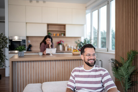 Happy man relaxing on sofa while woman using tablet in kitchen
