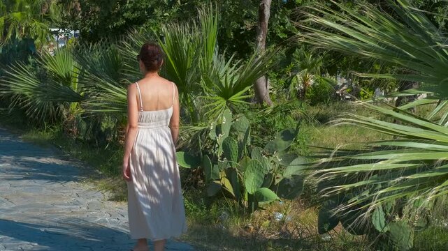 Woman walking away on a path in a tropical garden. Woman in a white dress walking away on a stone path in a lush tropical garden, surrounded by palm trees, surrounded by palm trees and other plants - Powered by Adobe