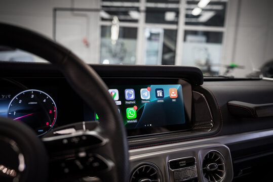 Close up of a luxury car dashboard featuring a digital display with app icons, circular metallic air vents, and carbon fiber trim in a showroom setting.