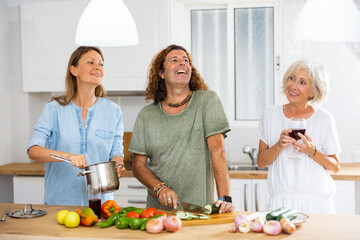 Happy curly man cooking vegetable salad while two excited women talking and drinking wine in the kitchen at home