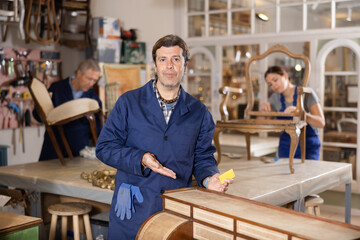 Professional middle-aged craftsman restorer in uniform renewing antique dresser while working in repair shop