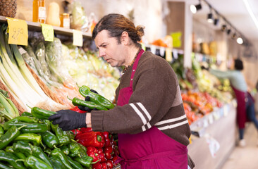 Adult man seller in apron puts fresh bell peppers on display in vegetable shop