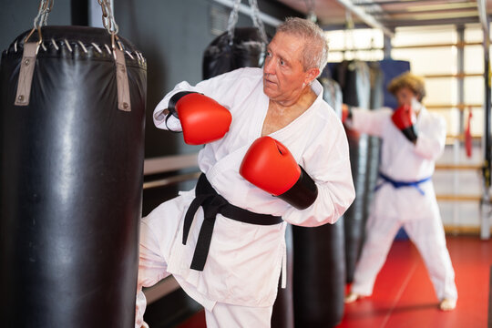 Senior man in white gi with black belt and red boxing gloves practicing kick on punching bag in martial arts dojo, staying active and focused - Powered by Adobe