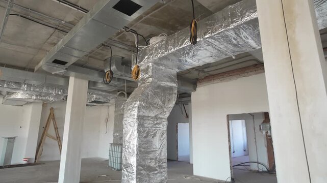 Air ventilation duct wrapped with silver foil and electrical cables coils in workshop. Unfinished industrial building with utilities