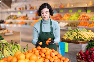 Shop seller puts goods on display case. Woman creates pyramid of tangerine, presents fresh goods. Vegetable shop near house. Spacious store with long rows of display cases, customers waiting