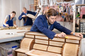 Skilled middle-aged male furniture repair specialist in uniform restoring Victorian dresser during workday in workshop