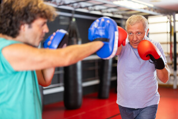 Concentrated mature hobby boxer in boxing gloves practicing self-defense techniques in gym, throwing punches on focus mitts in hands of instructor..