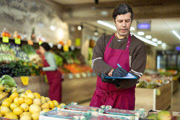 Adult man seller in apron conducting inventory with checklist in vegetable shop