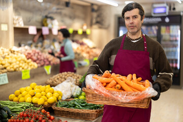 Man in apron carries large quantity of carrot in wicker basket to sales area, supplementing stores assortment with fresh vegetables