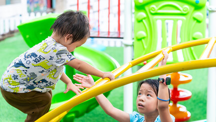 Fototapeta premium Kid exercise for health and sport concept. Children playing together on a playground in the summer sun. Happy Asian little child boy playing and climb on a steel arch bridge. My sister's concern.