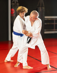 Experienced motivated senior man working on martial arts skills in training fight on red tatami...