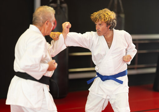 In modern dojo with red flooring and hanging punching bags, focused middle‑aged blue‑belt karateka in white gi working on strike and block combinations with older sparring partner - Powered by Adobe
