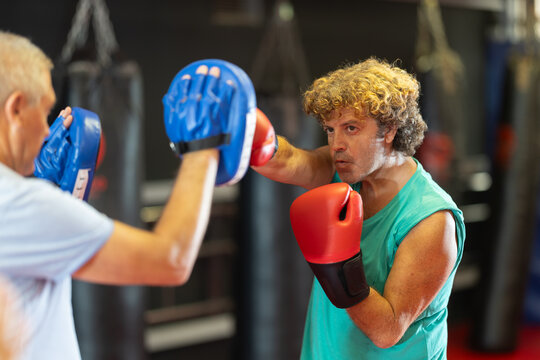 Focused middle-aged man wearing boxing gloves working out with older male partner during self-defense course, practicing punches on mitts.. - Powered by Adobe