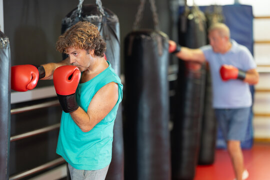 Motivated middle-aged man in everyday attire practicing striking hanging bag, refining practical techniques in group self-defense session at gym.. - Powered by Adobe