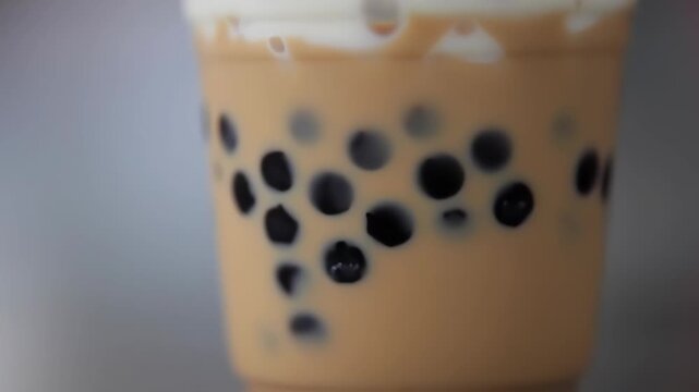 Close-up of a plastic cup filled with a light beige-tan drink, topped with a layer of off-white foam.  Dark tapioca balls are dispersed throughout the drink