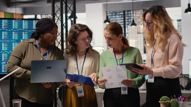 Ambitious and goal oriented diverse businesswomen reviewing corporate reports and graphs, working in synergy to achieve professional growth in a modern work environment. Camera B.