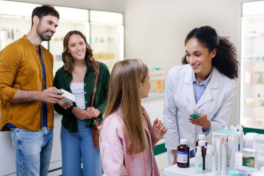 Professional friendly female pharmacist giving pills to preteen girl in modern drugstore, parents standing on background and smiling - Powered by Adobe