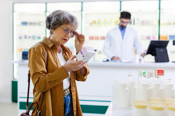 Naklejka premium Senior woman choosing to buy medicine or vitamins, reading information on package, pharmacist standing at counter on background. Pharmacy full of health care products