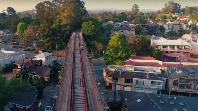 Two people walk along the historic trestle bridge in Capitola, California, USA. The bridge spans a residential area with lush trees and colorful houses.