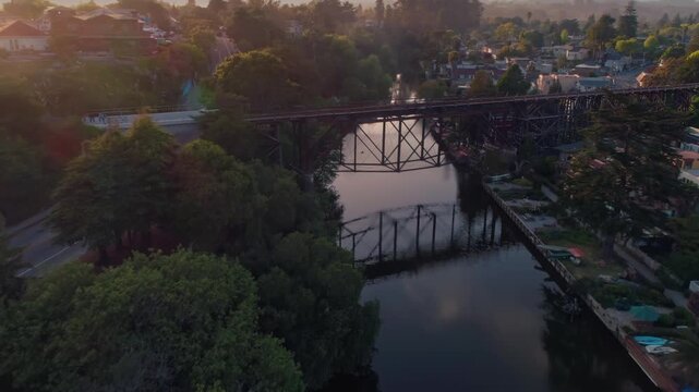 Aerial view of Capitola, California, USA, at sunset. Cars and pedestrians cross the bridge over the river, connecting the town. Houses line the riverbanks and hills.