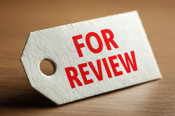 Close up of a white tag with the words for review in red lettering on a wood surface texture