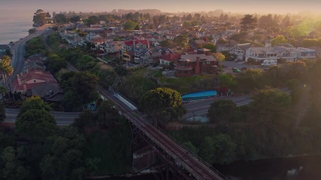 Aerial view of a coastal town in California, USA, featuring a railway bridge spanning a river, with houses and the Pacific Ocean in the background.
