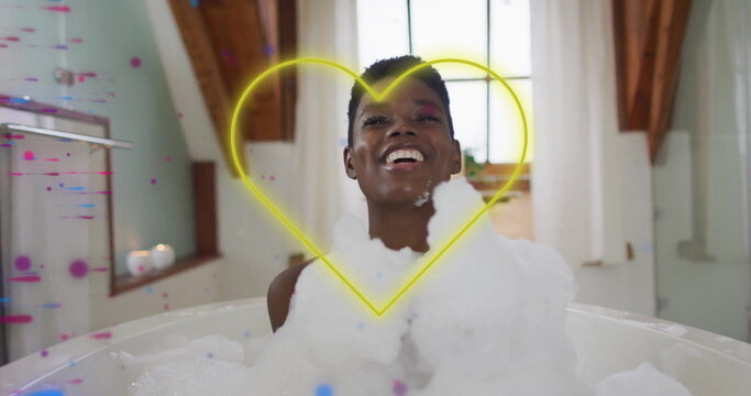 Laughing woman sitting in foam tub in bathroom, with neon heart overlay pixel effects and candles - Powered by Adobe